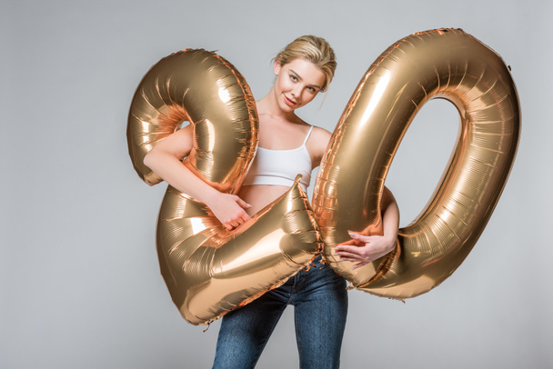 beautiful slim girl in jeans and white bra posing with 20 golden balloons, isolated on grey - Foto, Imagem