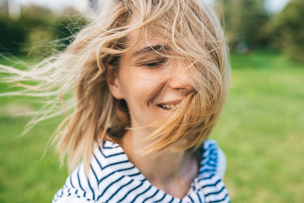 Cropped close up outdoor shot of attractive happy female with blond blowing hair and closed eyes enjoy weather. Close up shot of beautiful young woman smiling with windy hair in park. People concept - Foto, Imagen
