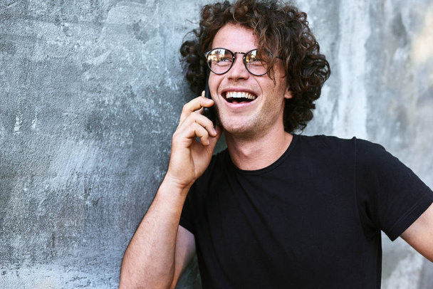 laughing, happy handsome man with curly hair talking on mobile phone while standing against grey wall - Φωτογραφία, εικόνα