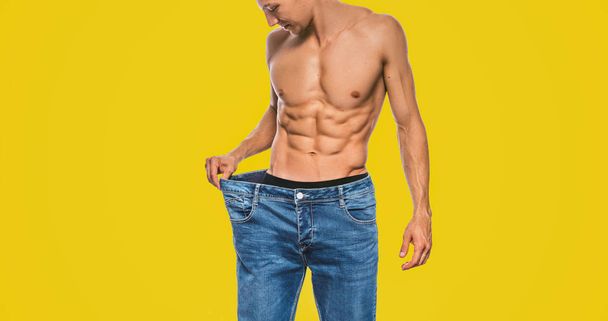Horizontal studio image of young Caucasian fit man wearing big blue jeans after diet, showing sexy torso. Attractive man posing on yellow background after losing weight. Copy space. Healthy lifestyle - Photo, Image