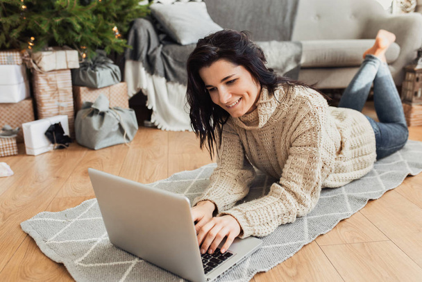 Image of beautiful smiling young woman shopping online on laptop in cozy Christmas interior. Female lying on the floor next the Christmas tree and sofa, bying gifts for the family on winter sales. - Фото, зображення
