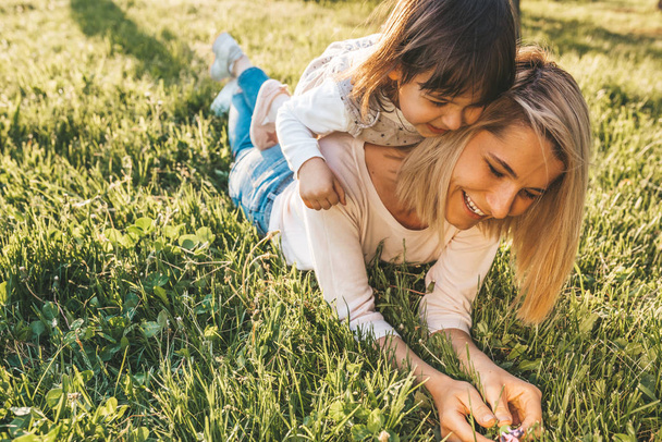 Attractive happy young mother lying with her cute daughter on grass in the park. Happy family relationship. Cheerful woman playing with her child outdoor, explore nature. Motherhood and childhood.  - Photo, image