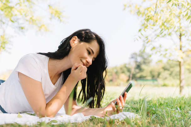 horizontales Porträt einer lächelnden jungen schönen Frau mit glücklichem Blick, während sie mit dem Handy telefoniert, gute Nachrichten empfängt und im Park auf weißer Decke und grünem Gras sitzt. Menschen und Lebensstil. - Foto, Bild