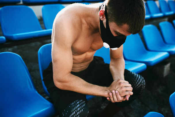 Side view of young muscular male relaxing and sitting on the seat at the stadium with mask after exercises. Portrait of shirtless sportsman relaxing after workout in stadium outside. People and sport - Φωτογραφία, εικόνα
