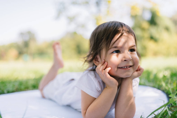 Horizontal portrait of happy smile toddler girl have fun outdoor in the park, sitting on a white blanket. Positive expression of a child. Adorable kid on nature sunlight. Childhood concept. - Foto, imagen