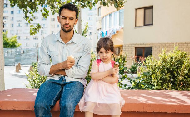 Unhappy cute little girl sitting with dad on the city street and eating ice-cream outdoor. Upset girl kid and sad father have negative emotions. Good relationship between dad and daughter - Photo, Image