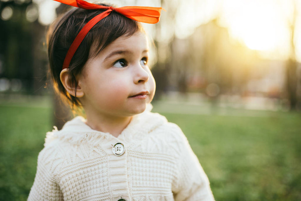 Cute pretty girl toddler with red ribbon on head, looking away, on green grass and sunset background. Caucasian happy baby smiling with a diffused green grass background. Childhood lifestyle. - Foto, imagen