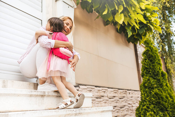 Horizontal image of happy mother with meets her pupil daughter going back at home from kindergarten. Cute little girl with mom embrace each other, feeling happy after the first preschool day. Family - Photo, Image