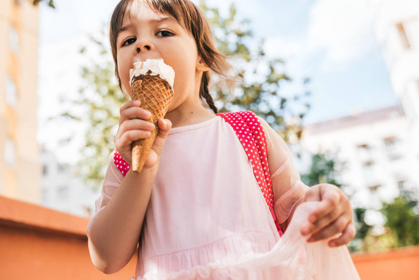 Horizontal closeup portrait of cute little girl walking along city street and eating ice cream outdoor. Happy kid girl wears pink dress eat not healthy snack. Happy preschooler with pink backpack. - Photo, Image