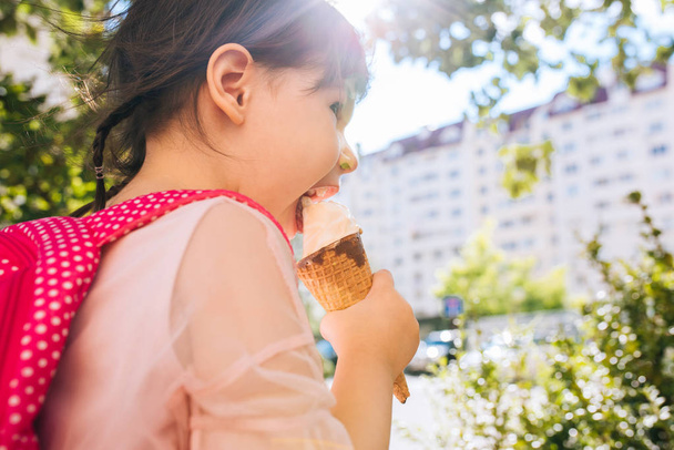 Rear view closeup portrait of cute little girl walking along city street and eating ice-cream outdoor. Happy kid girl wears pink dress tasting dessert after preschool. Happy toddler with pink backpack - Fotoğraf, Görsel