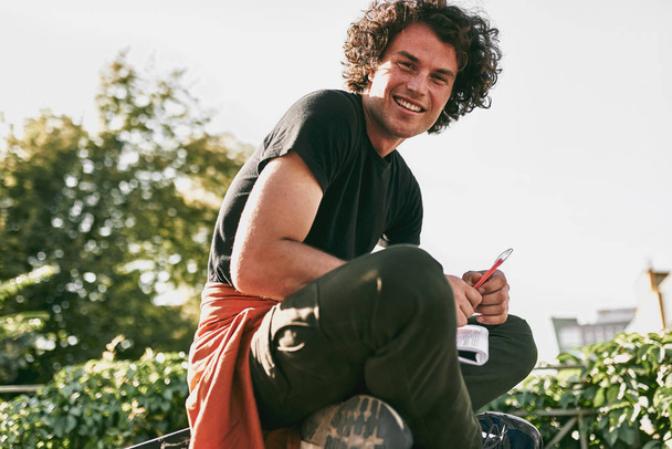 Side view of man student with curly hair, smiling and wearing black t-shirt sitting on the street and preparing for exams. Freelancer businessman planning the day sitting on the city street - Foto, Imagen