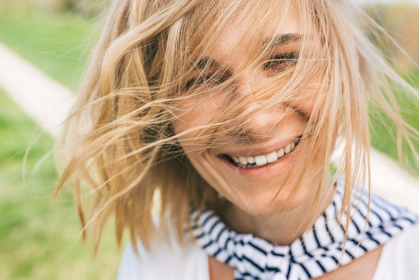Retrato close-up de alegre mulher feliz com cabelo loiro soprando, sorrindo com dente branco sorriso saudável e olhando para a câmera. Jovem sorrir com o cabelo ventoso no parque. Conceito de pessoas
 - Foto, Imagem