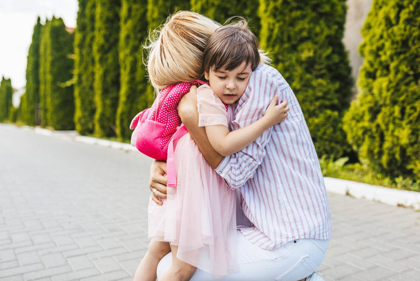 Horizontal image of beautiful mother with love hugging her cute daughter in the street next to the home. Good relationship support. Sad little girl embracing her mom before the preschool day - Foto, Imagen
