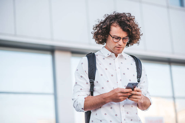 Stylish young serious Caucasian male wearing trendy spectacles and shirt looking at screen of his smart phone, about to answer to some email. People, lifestye and modern technology communication. - Фото, изображение