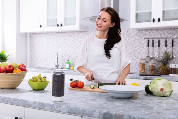 Happy Woman Talking To Voice Assistant While Slicing Cucumber With Kitchen Knife - Фото, зображення