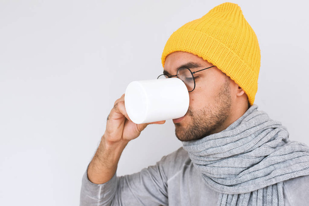 Side view of handsome male having sore throat influenza. Portrait of sick man have grippe and drinking cup of tea, wearing glasses, yellow hat, grey sweater and scarf. People, health, medicine concept - Foto, Bild