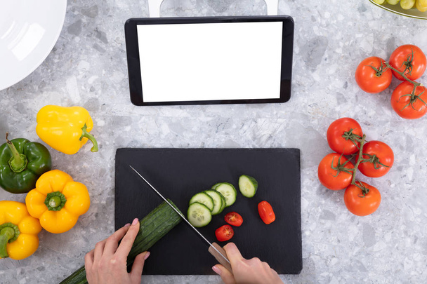 Woman Cutting Cucumber With Kitchen Knife Near Digital Tablet, Tomatoes And Bell Peppers On Kitchen Counter - Фото, изображение