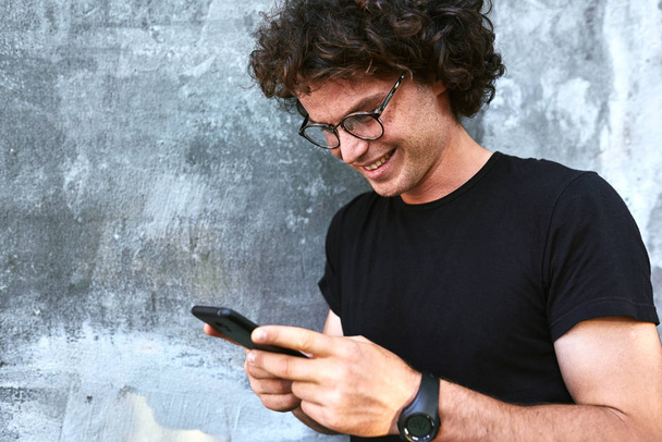smiling handsome man with curly hair holding mobile phone while standing against grey wall - Zdjęcie, obraz