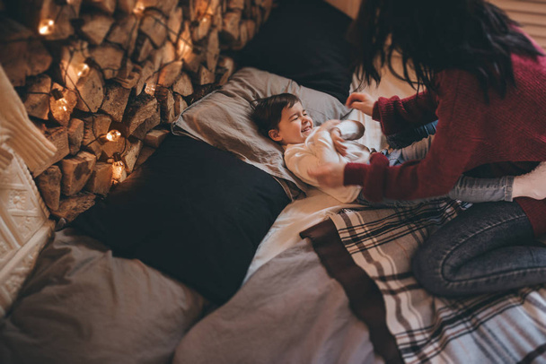 Above shot of happy mother and her little daughter playing on the bed with cozy and christmas lights decoration. Beautiful woman and cute little girl have fun in winter weekend. Family, people, xmas - Φωτογραφία, εικόνα