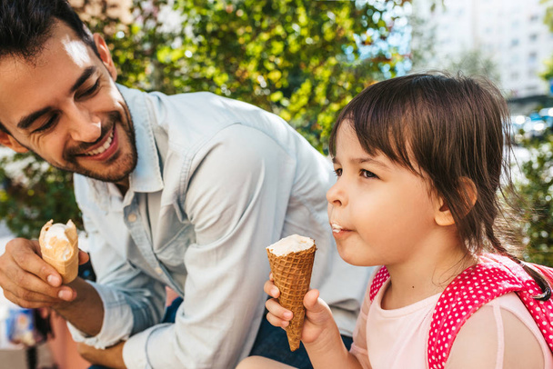 Closeup shot of happy cute little girl sitting with handsome dad on the city street and eating ice-cream outdoor. Fun girl kid and father have fun outside. Good relationship between dad and daughter - Foto, Imagem
