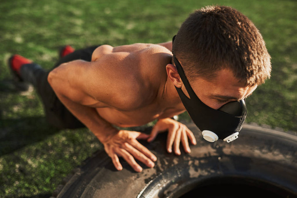 Closeup portrait of Caucasian fitness muscular male in mask to increase load on breathing muscles doing push-ups exercises on tire outdoors. Copy space. Shirtless sportsman doing workout on stadium. - Φωτογραφία, εικόνα