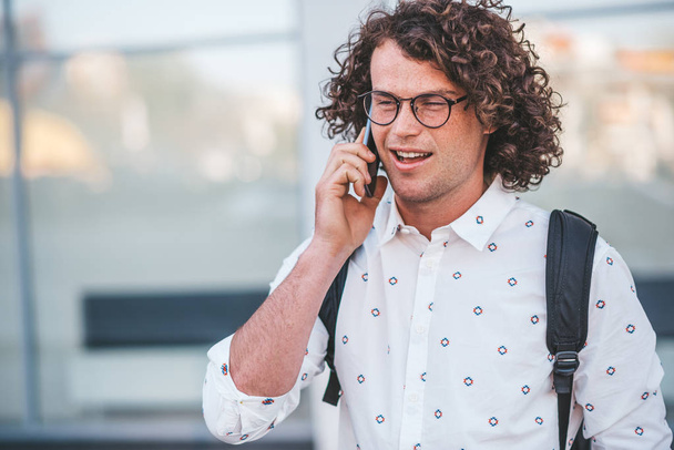 Portrait of stylish curly hair male in trendy spectacles standing outdoors while holding backpack and talking by his smart phone with his girlfriend on the city street. People, technology, lifestyle.  - Фото, изображение
