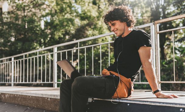 Side view image of cheerful Caucasian male with curly hair using laptop computer and smiling while sitting on the pavement. Young freelance business man working outside.  - Photo, image