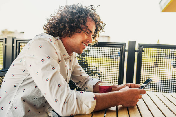 Closeup side view portrait of smiling handsome male with curly hair in stylish shirt, browsing on free wifi via smart phone.Attractive man texting message on cafe outdoor.People, lifestyle, technology - Photo, Image