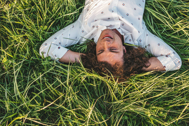 top view of happy Caucasian young man with curly hair smiling and lying on green grass in park - Foto, imagen