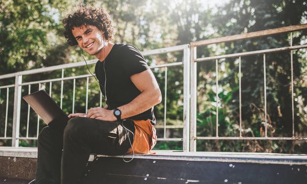 Horizontal image of cheerful Caucasian male using laptop computer and smiling while sitting on the pavement. Young freelance business man working outside.  - 写真・画像