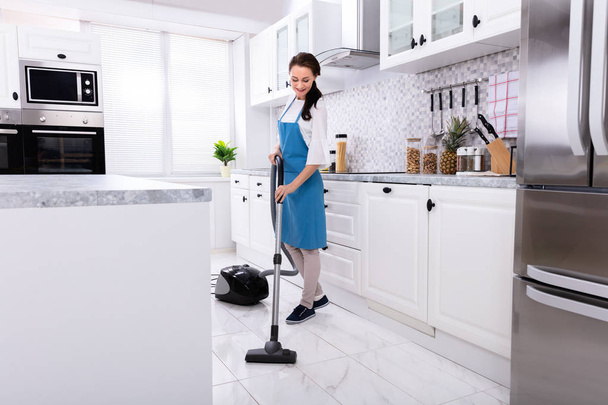 Young Female Janitor In Uniform Cleaning Kitchen Floor With Vacuum Floor - Zdjęcie, obraz