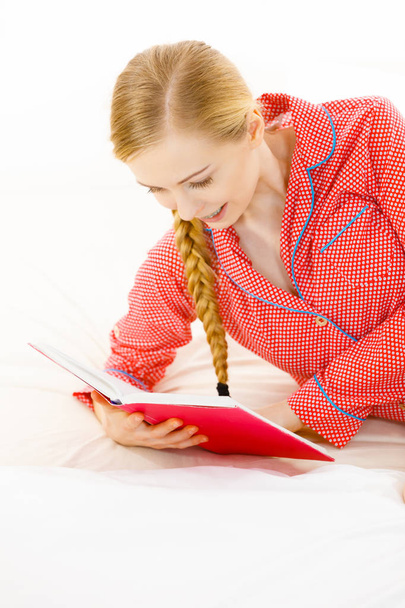 Girl lying in bed reading book. Young blonde female wearing red dotted pajamas relaxing at home on mattress. - Φωτογραφία, εικόνα