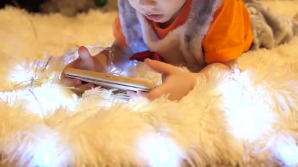 L'enfant repose sur une couverture blanche douce dans la chambre des enfants. Il regarde des dessins animés sur le smartphone. Guirlande de Noël. Bonne enfance
 - Séquence, vidéo