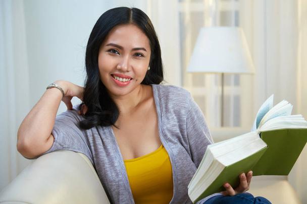 Senhora filipina bonita sorrindo e olhando para a câmera enquanto sentado em sofá confortável na sala de estar aconchegante e lendo livro interessante
 - Foto, Imagem