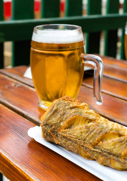 Smažená ryba na jednorázové papírové desce a chlazené sklo návrhu piva na dřevěném stole v venkovní kavárně. Selektivní zaměření - Fotografie, Obrázek