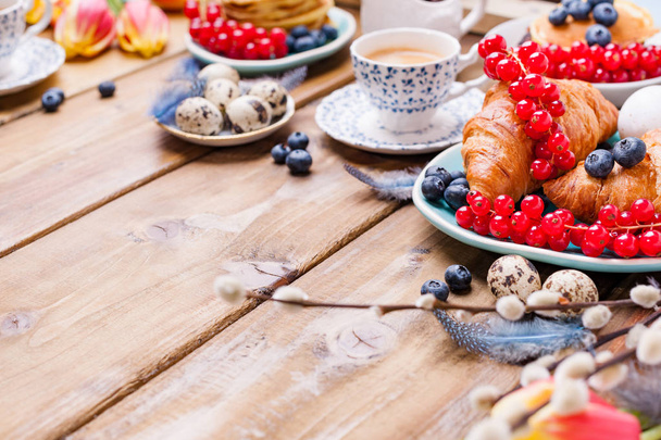 Easter breakfast flat lay with fresh coffee, berries and pastries , orange tulips, croissants with bacon and various sweets , colored quail eggs and spring holidays decorations. Top view. Copy space - Foto, immagini