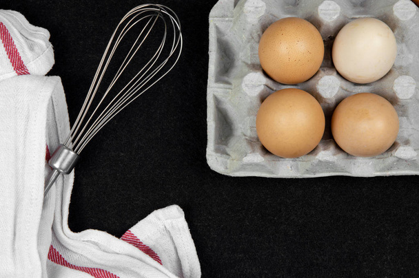 Vista superior de ovos de galinha em uma caixa de papelão e um batedor de ovos em um fundo preto
. - Foto, Imagem