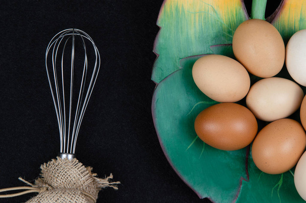 Vista superior de ovos de galinha em uma placa em forma de folha e um batedor de ovos em um fundo preto
. - Foto, Imagem