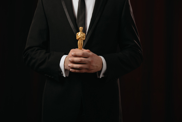 KYIV, UKRAINE - JANUARY 10, 2019: cropped view of man in suit holding oscar award on dark background - Foto, afbeelding