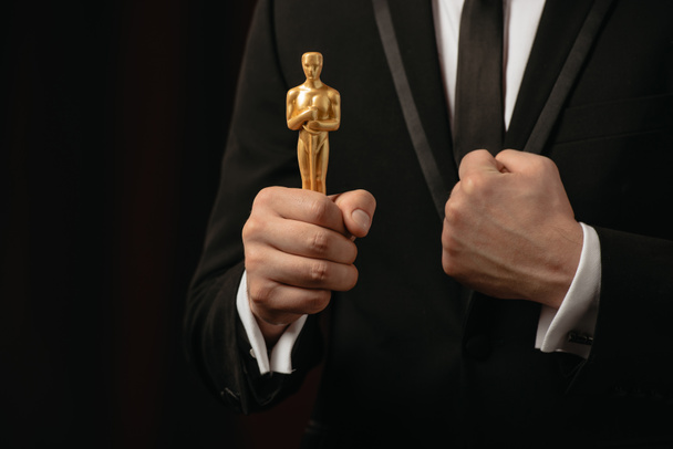 KYIV, UKRAINE - JANUARY 10, 2019: partial view of man in suit with clenched fist holding oscar award isolated on black - Foto, imagen