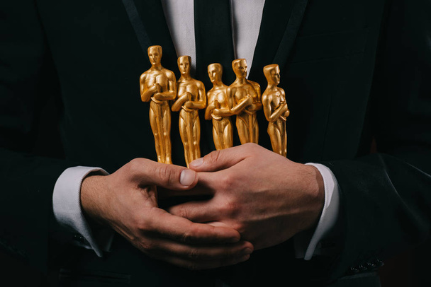 KYIV, UKRAINE - JANUARY 10, 2019: partial view of man in suit holding oscar awards isolated on black - 写真・画像