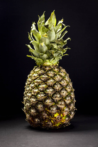 Close-up shot of ripe yellow pineapple with textured peel and green leaves on black background - 写真・画像