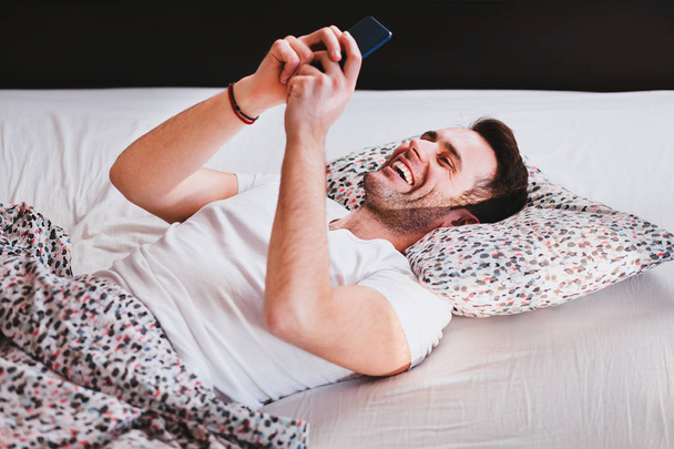 Jonge man met smartphone in bed in de ochtend - Foto, afbeelding