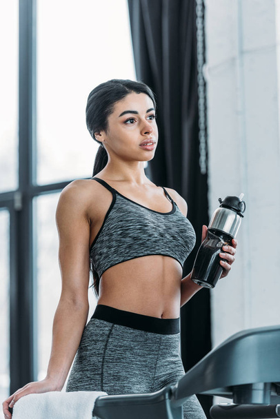 pensive young african american woman standing on treadmill and holding sport bottle - Foto, Bild