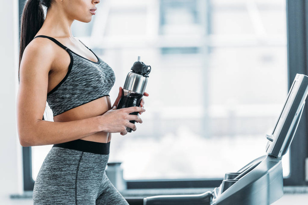 cropped shot of sporty african american girl holding sports bottle and training on treadmill in gym - Zdjęcie, obraz