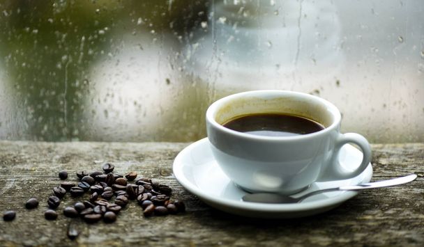 Autumn cloudy weather better with caffeine drink. Enjoying coffee on rainy day. Coffee morning ritual. Fresh brewed coffee white mug and beans on windowsill. Wet glass window and cup of hot coffee - Zdjęcie, obraz