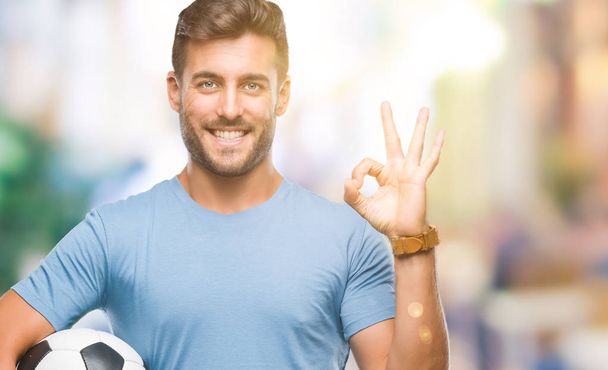 Όμορφος νεαρός, κρατώντας την μπάλα ποδοσφαίρου ποδοσφαίρου πάνω από απομονωμένες φόντο κάνει εντάξει πινακίδα με τα δάχτυλα, εξαιρετική σύμβολο - Φωτογραφία, εικόνα