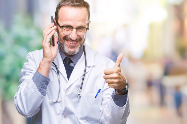 Старший седовласый врач средних лет разговаривает по смартфону на изолированном фоне счастливый с большой улыбкой делает хорошо знак, палец вверх пальцами, отличный знак
 - Фото, изображение