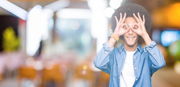 Young african american man with afro hair doing ok gesture like binoculars sticking tongue out, eyes looking through fingers. Crazy expression. - Photo, Image