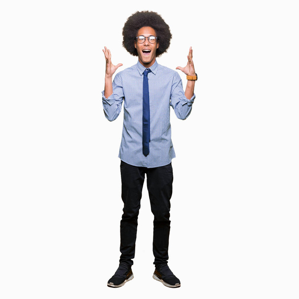 Joven hombre de negocios afroamericano con el pelo afro usando gafas celebrando loco y sorprendido por el éxito con los brazos levantados y los ojos abiertos gritando emocionado. Concepto ganador
 - Foto, imagen